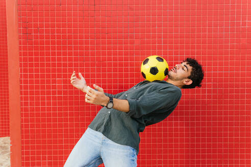 Seitenansicht eines jungen hispanischen Mannes in schwarzem Hemd, der einen gelben Fußball auf der Brust gegen eine rote Kachelwand auf der Straße balanciert - ADSF39810