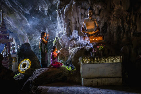 Ein Höhlentempel mit Buddha-Statuen, brennenden Kerzen und zwei knienden, betenden Mädchen. - MINF16565