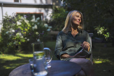 Ältere Frau mit Buch sitzt auf einem Stuhl im Garten - RIBF01118