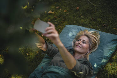 Reife Frau liegt auf Gras und nimmt Selfie durch Smartphone im Garten - RIBF01165