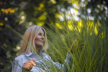 Reife Frau mit blondem Haar berührt Pflanze im Garten - RIBF01181