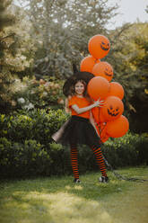 Lächelndes Mädchen mit Hexenhut hält orangefarbene Luftballons im Hinterhof - MDOF00103