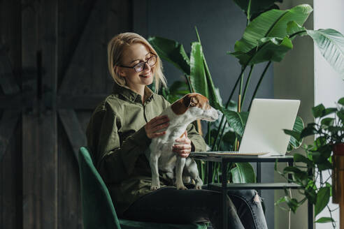 Lächelnde Geschäftsfrau, die einen Hund streichelt, der auf einem Stuhl im Büro zu Hause sitzt - VSNF00052