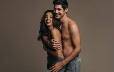 Fröhliches Paar, das nur Jeans trägt, steht zusammen auf braunem Hintergrund. Lächelnde Frau wird von ihrem Freund von hinten umarmt. - JLPSF11372