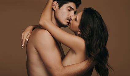 Zärtliches junges Paar, das sich küsst. Mann und Frau oben ohne küssen sich leidenschaftlich auf braunem Hintergrund. - JLPSF11383