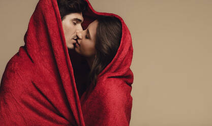Liebendes junges Paar, das sich unter einer roten Decke küsst. Junger Mann und Frau in eine Decke gewickelt und küssend auf braunem Hintergrund. - JLPSF11397