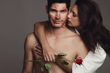 Girl holding a rose embracing her man from behind and kissing with lipstick marks left on the face. Romantic young couple on grey background. - JLPSF11411