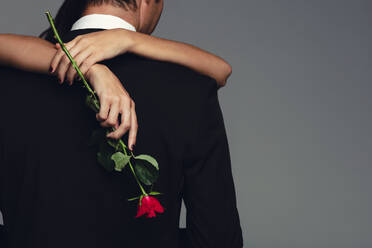 Rückansicht einer Frau, die eine Rose hält und einen Mann im schwarzen Anzug umarmt. Ein liebendes Paar, das sich auf grauem Hintergrund umarmt. - JLPSF11435
