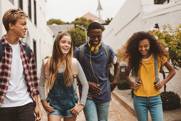 Gruppe multiethnischer College-Freunde, die auf der Straße spazieren gehen. Lächelnde Teenager-Freunde mit College-Taschen, die im Freien spazieren gehen und sich unterhalten. - JLPSF11440