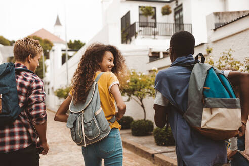 Rückansicht von Teenager-Freunden, die zusammen auf der Straße gehen. Lächelndes Teenager-Mädchen, das mit Freunden spazieren geht und eine College-Tasche trägt. - JLPSF11441