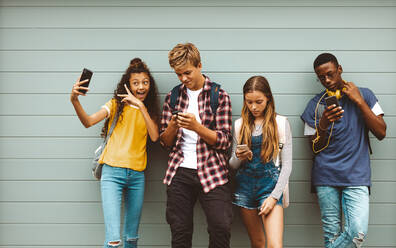 College friends standing outdoors looking using their cell phones. Young girl taking a selfie standing outdoors against a wall with her friends looking at their mobile phones. - JLPSF11448