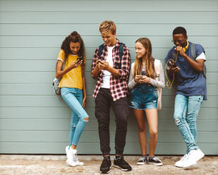 Gruppe multiethnischer Freunde, die auf der Straße stehen und auf ihre Handys schauen. Glückliche Teenager-Jungs und -Mädchen, die College-Taschen tragen und sich unterhalten, während sie auf ihre Handys schauen. - JLPSF11449