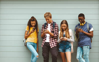 College friends wearing bags using mobile phones standing outdoors. Multiethnic teenage boys and girls standing outdoors against a wall and looking at their mobile phones. - JLPSF11450