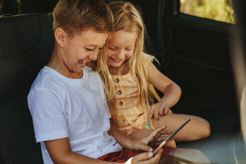 Zwei Kinder sitzen auf der Rückbank eines Autos und spielen Spiele auf einem digitalen Tablet. Sie sind mit dem Auto unterwegs. - JLPSF11459