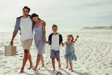 Beautiful family of four on summer vacation at beach. Family walking together on beach, with man carrying a picnic basket. - JLPSF11462