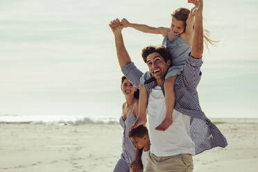 Family of four on a beach vacation. Father giving daughter ride on shoulders as they walk at the seashore with mother and son. - JLPSF11465