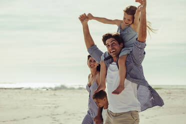 Vierköpfige Familie im Strandurlaub: Vater lässt Tochter auf den Schultern reiten, während sie mit Mutter und Sohn am Meer spazieren gehen. - JLPSF11465