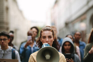 Junge Frau mit einem Megaphon und einer Gruppe von Demonstranten im Hintergrund, die mit einem Megaphon in der Stadt protestieren. - JLPSF11512