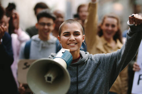 Young woman standing outdoors with group of demonstrator in background. Woman protesting with a megaphone outdoors on road. - JLPSF11516