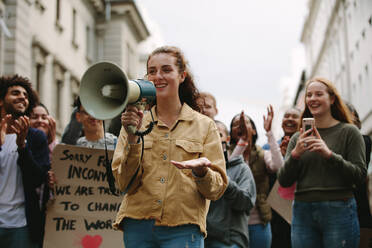 Frau mit Megaphon bei einer Kundgebung im Freien auf der Straße. Frau steht im Freien mit einer Gruppe von Demonstranten, die klatschen und feiern. - JLPSF11570