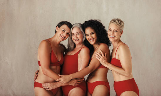 Aufnahme von lächelnden Frauen, die ihre natürlichen und alternden Körper umarmen. Vier glückliche und körperbewusste Frauen jeden Alters, die sich gegenseitig umarmen, während sie rote Unterwäsche vor einem Studiohintergrund tragen. - JLPSF11595
