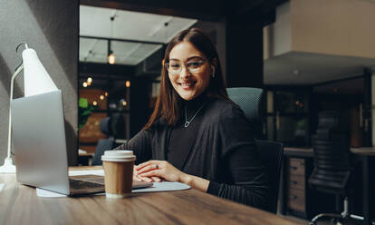 Cheerful young businesswoman smiling at the camera while working in a modern co-working office. Happy female entrepreneur using wireless earphones and a laptop while working remotely. - JLPSF11615