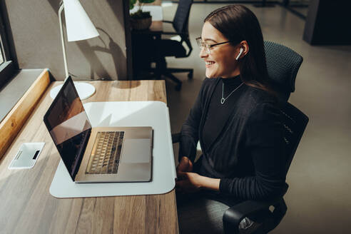 Glückliche junge Geschäftsfrau, die während eines virtuellen Meetings in einem modernen Co-Working-Büro lächelt. Fröhliche Unternehmerin, die mit ihren Geschäftspartnern kommuniziert, während sie aus der Ferne arbeitet. - JLPSF11617