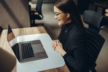 Weibliche Geschäftsfrau, die an einem Online-Meeting in einem modernen Co-Working-Büro teilnimmt. Glückliche junge Geschäftsfrau, die mit ihren Geschäftspartnern kommuniziert, während sie aus der Ferne arbeitet. - JLPSF11618