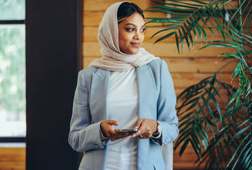 Muslimische Geschäftsfrau, die nachdenklich wegschaut, während sie ein Smartphone in der Hand hält. Junge Geschäftsfrau, die in einem Büro eine Textnachricht sendet. Unternehmerin, die an einem modernen Arbeitsplatz einen Hijab trägt. - JLPSF11671