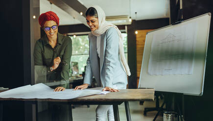 Cheerful female architects working on blueprint drawings in a modern office. Two Muslim businesswomen planning a new innovative project. Creative designers wearing headscarfs in an inclusive workplace. - JLPSF11677