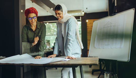 Fröhliche Architektinnen, die in einem modernen Büro an Entwürfen arbeiten. Zwei muslimische Geschäftsfrauen, die ein neues innovatives Projekt planen. Kreative Designerinnen mit Kopftuch an einem integrativen Arbeitsplatz. - JLPSF11677