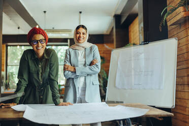 Happy Muslim designers smiling at the camera while working on blueprint drawings. Two creative businesswomen planning a new project in an office. Architects wearing headscarfs in a modern workplace. - JLPSF11678