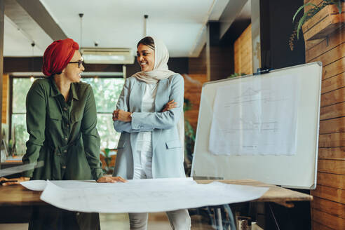 Fröhliche Designerinnen diskutieren bei der Arbeit an Bauplänen. Zwei kreative Geschäftsfrauen planen ein neues Projekt in einem Büro. Architektinnen mit Kopftüchern an einem modernen Arbeitsplatz. - JLPSF11679