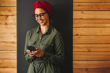 Muslimische Geschäftsfrau, die ihr Smartphone benutzt, während sie an einer Wand steht. Fröhliche Geschäftsfrau, die lächelt, während sie eine Textnachricht sendet. Unternehmerin mit Kopftuch an einem modernen Arbeitsplatz. - JLPSF11697