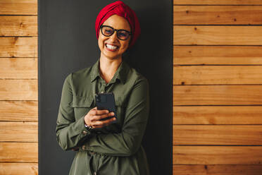 Glückliche muslimische Unternehmerin, die in die Kamera lächelt und ein Smartphone in der Hand hält. Fröhliche Geschäftsfrau, die in einem Büro eine Textnachricht liest. Unternehmerin mit Kopftuch an einem modernen Arbeitsplatz. - JLPSF11698