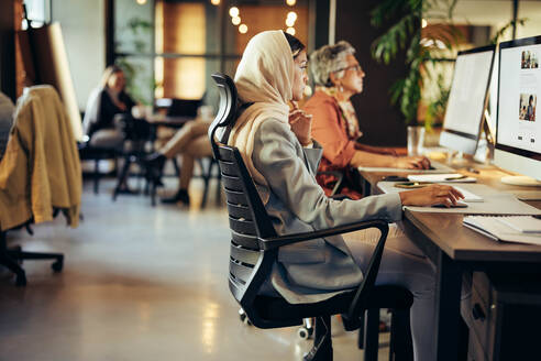 Eine muslimische Geschäftsfrau arbeitet an einem Computer in einem modernen Büro. Eine kreative Designerin trägt einen Hijab an einem integrativen Arbeitsplatz. - JLPSF11721