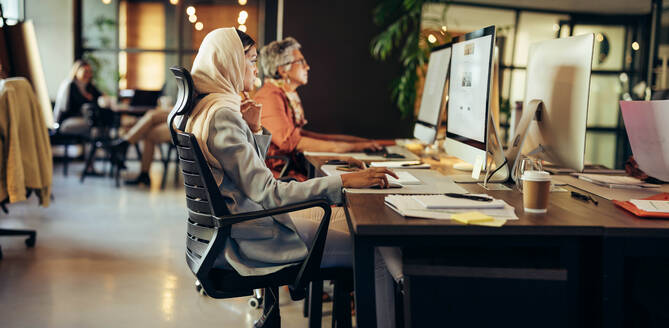 Kreative Geschäftsfrau bei der Arbeit am Computer in einem modernen Co-Working-Büro. Muslimische Designerin trägt einen Hijab an einem integrativen Arbeitsplatz. - JLPSF11722