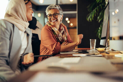 Zwei glückliche Geschäftsfrauen diskutieren an ihren Schreibtischen in einem Büro. Verschiedene Geschäftsfrauen arbeiten als Team in einem kreativen Co-Working-Büro. - JLPSF11723