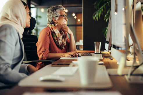 Zwei Geschäftsfrauen lächeln fröhlich, während sie in einem Büro auf einen Computerbildschirm schauen. Verschiedene Geschäftsfrauen arbeiten als Team in einem kreativen Co-Working-Büro. - JLPSF11726