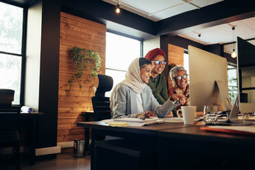 Cheerful businesswomen working as a team in a modern office. Group of multicultural businesswomen smiling cheerfully while looking at a computer screen in an inclusive workplace. - JLPSF11730