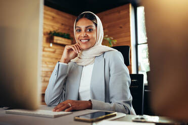 Glückliche muslimische Geschäftsfrau, die in die Kamera lächelt, während sie in einem modernen Büro an einem Computer arbeitet. Fröhliche junge Geschäftsfrau, die einen Hijab an einem integrativen Arbeitsplatz trägt. - JLPSF11733