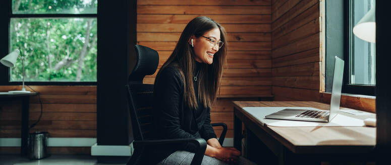 Lächelnde Geschäftsfrau bei einem Online-Meeting in einem modernen Co-Working Space. Glückliche Unternehmerin, die mit ihren Geschäftspartnern kommuniziert, während sie aus der Ferne arbeitet. - JLPSF11736
