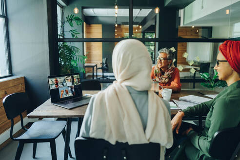 Eine Gruppe ethnischer Geschäftsfrauen nimmt an einem Online-Meeting in einem modernen Büro teil. Multikulturelle Geschäftsfrauen führen eine Videokonferenz mit ihren Geschäftspartnern an einem kreativen Arbeitsplatz durch. - JLPSF11762