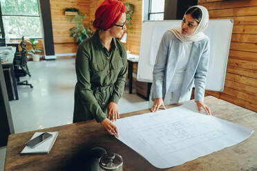 Muslimische Designerinnen im Gespräch bei der Arbeit an einem Bauplan. Zwei kreative Geschäftsfrauen planen ein innovatives Projekt. Architektinnen mit Kopftuch an einem integrativen Arbeitsplatz. - JLPSF11772