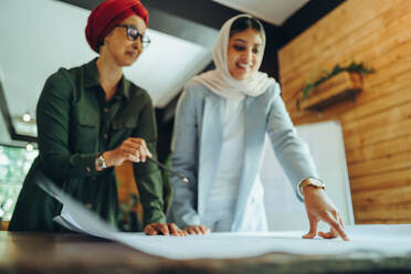 Female architects working on blueprint drawings in a modern office. Two Muslim businesswomen planning a new innovative project. Creative designers wearing headscarfs in an inclusive workplace. - JLPSF11778