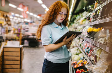 Frau, die in einem Supermarkt arbeitet. Weibliche Assistentin, die ein digitales Tablet benutzt, um eine Bestandsaufnahme der Produkte in einem Regal in einem Lebensmittelgeschäft zu machen. - JLPSF11828