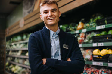 Grocery store assistant standing with his arms crossed. Happy young man working at supermarket. - JLPSF11832