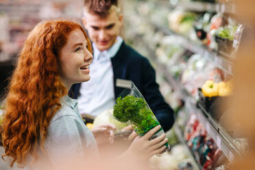 Zwei Personen, die in der Obst- und Gemüseabteilung eines Supermarktes arbeiten. Ein Mann und eine Frau, die in einem Supermarkt eine Ausbildung absolvieren, halten frisches Gemüse in der Hand, während sie die Gänge auffüllen. - JLPSF11834