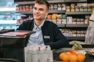 Man working at cash counter of a supermarket. Teenage cashier working at grocery store checkout. - JLPSF11837