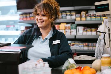 Happy woman cashier at a supermarket attending customer. Woman working at grocery store checkout talking with customer and smiling. - JLPSF11841